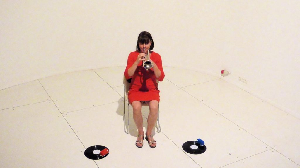 Eine Frau im roten Kleid sitzt in einem weißen Raum und spielt Trompete. Rechts und links von ihr liegt je eine Schallplatte auf dem Boden, darauf ein roter bzw. blauer Spielzeugplattenspieler in Form eines VW-Bullis.