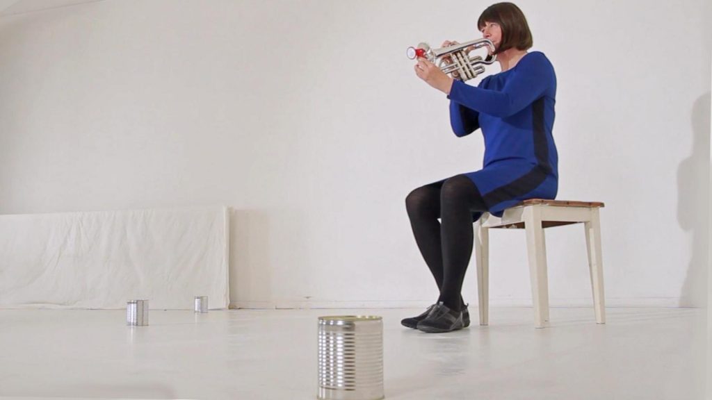 Eine Frau im blauschwarzen Kleid sitzt auf einem Hocker in einem weißen Raum und spielt Trompete. Auf dem Boden stehen drei Blechdosen im Raum verteilt.